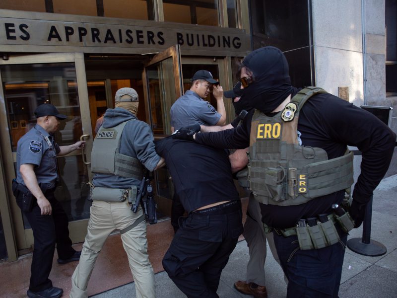 Law enforcement officers escort a handcuffed individual into a building labeled "APPRAISERS BUILDING" as other officers stand nearby.