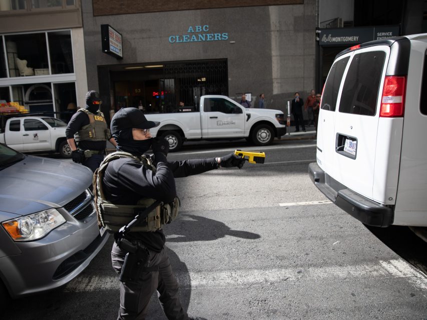A law enforcement officer in tactical gear points a yellow Taser at a white van on a city street near an ABC Cleaners storefront.