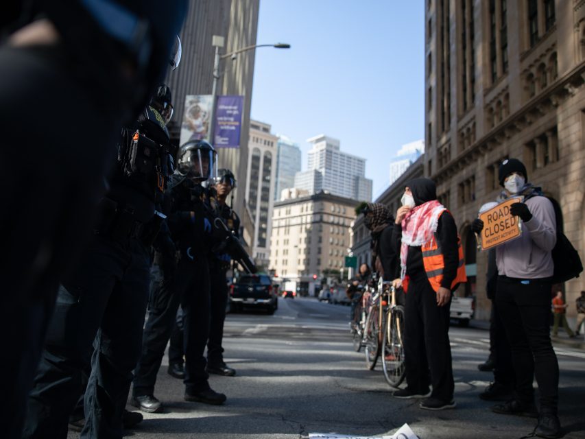 A group of police officers face protesters, some with bicycles; one protester holds a "Road Closed" sign on a city street with tall buildings in the background.