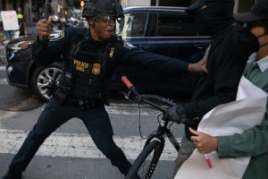 A police officer wearing protective gear confronts a masked individual holding a bicycle during a street incident. Another person stands nearby holding a sign or poster.