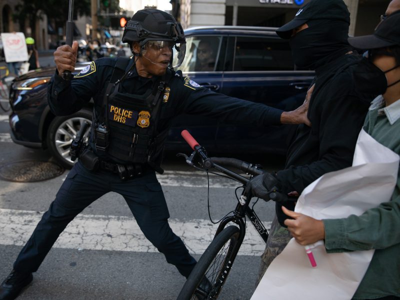 A police officer wearing protective gear confronts a masked individual holding a bicycle during a street incident. Another person stands nearby holding a sign or poster.