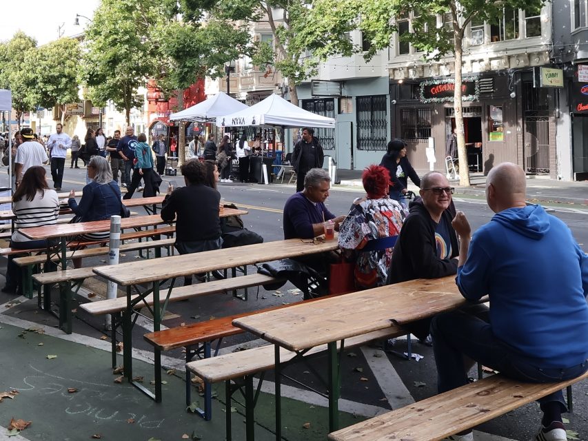People sit at wooden tables on a closed-off street with vendor tents, others walk or stand nearby; trees and buildings line the background.