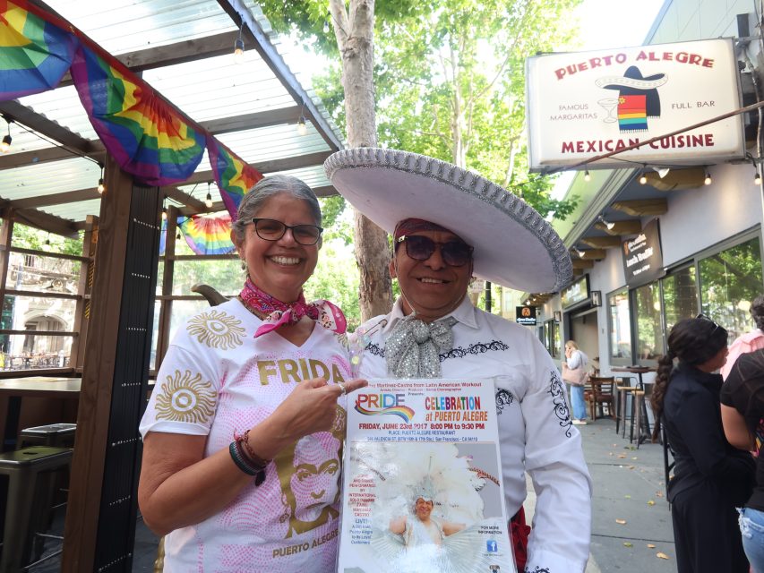 Two people in festive attire stand outside Puerto Alegre Mexican Cuisine, holding a Pride celebration flyer. Rainbow flags and restaurant signs are visible in the background.