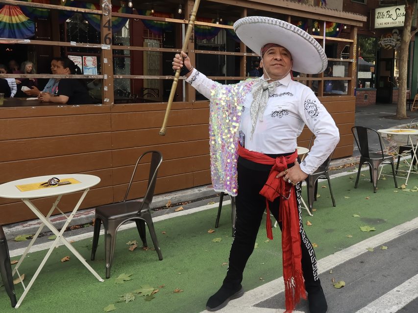 A person in a white mariachi outfit and sombrero stands on a city street, holding a staff and draped in a sequined cape near outdoor café tables.