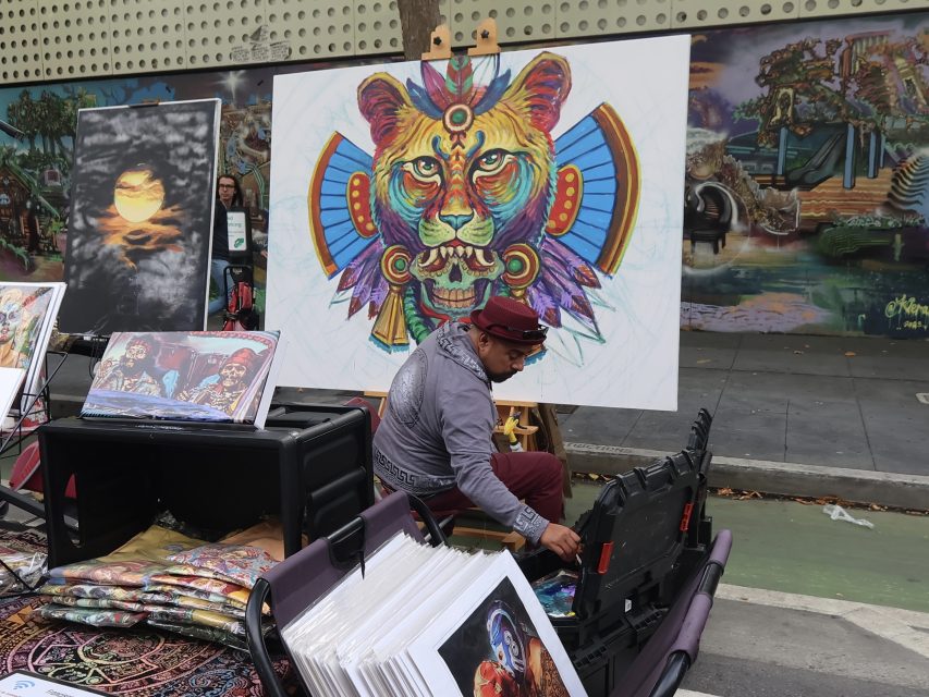 An artist sits by a display of colorful artworks and prints at an outdoor market, with a large painting of a stylized lion head prominently featured behind him.
