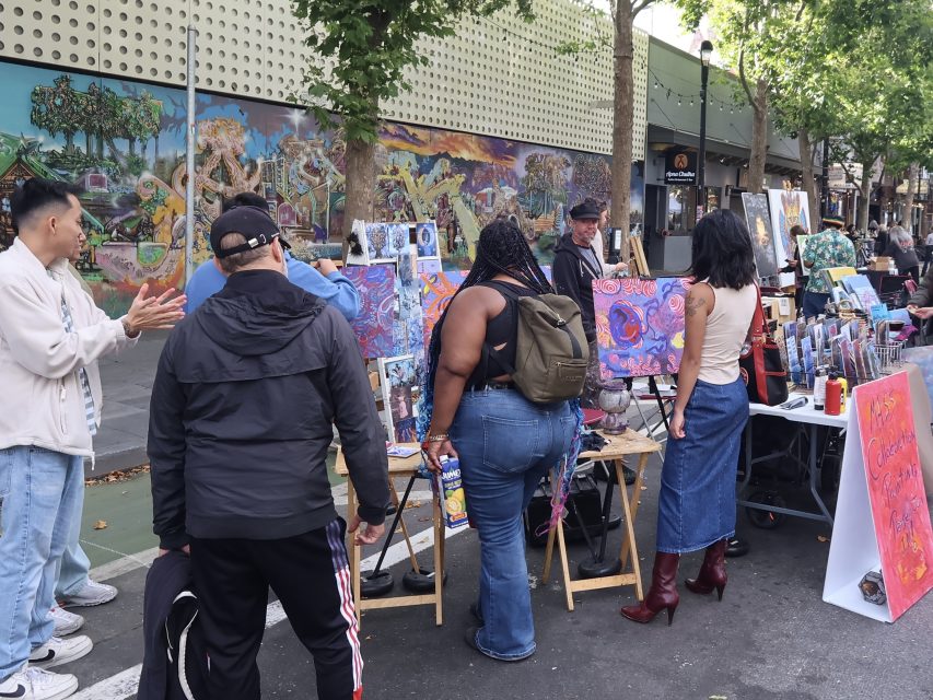 People gather around outdoor tables displaying colorful paintings at an art fair on a city street lined with murals and trees.