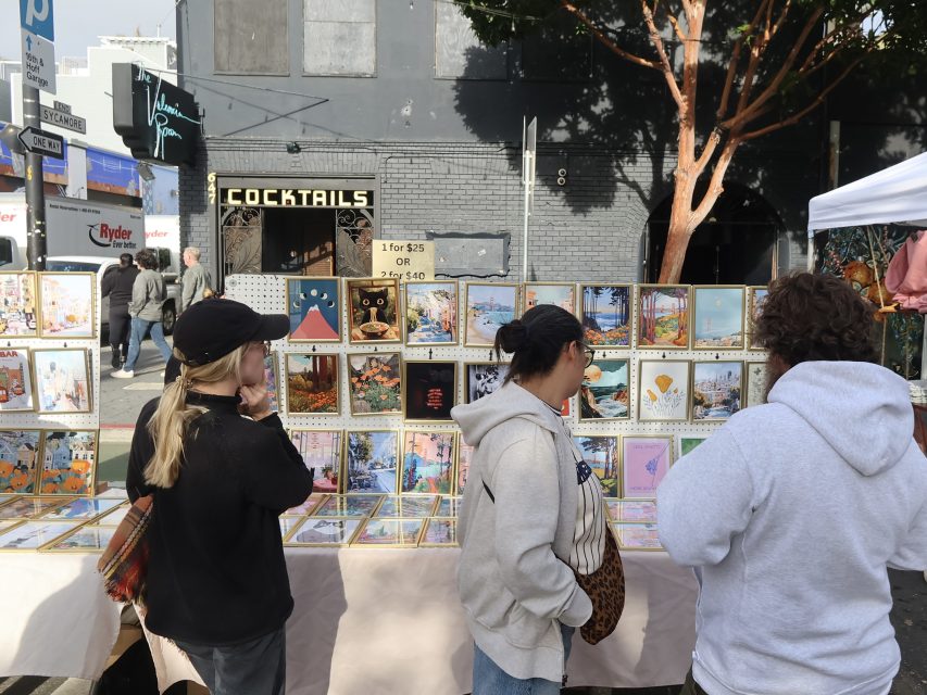 Three people look at framed artwork displayed on a table at an outdoor market, with a gray building and a "Cocktails" sign in the background.