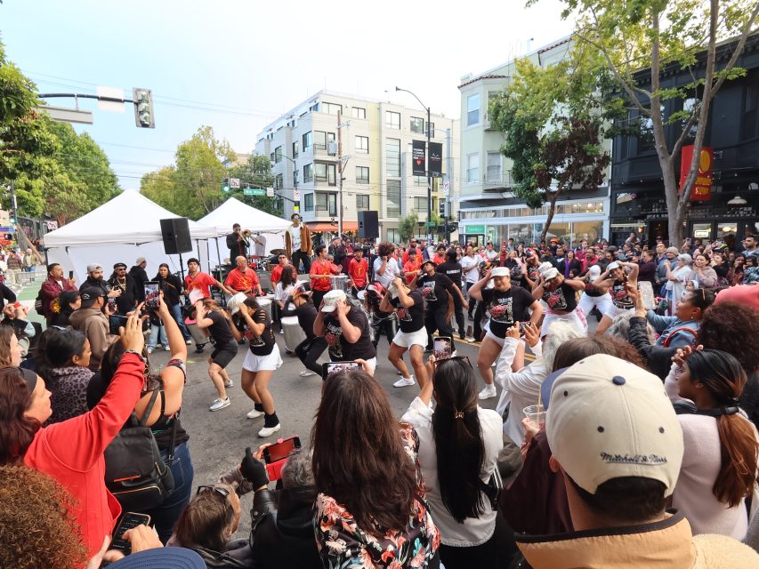 A large crowd watches a street performance featuring a dance group in coordinated outfits, with musicians in red jackets performing on a stage in the background.