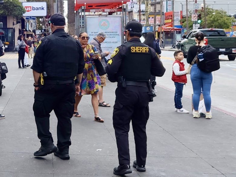 Two sheriff officers stand on a busy city sidewalk while pedestrians, including children and adults, walk by and interact.