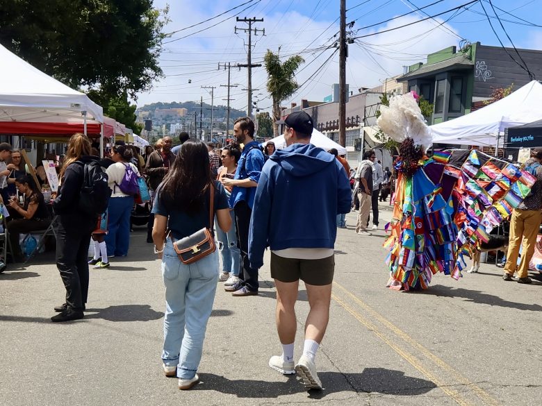 People walk through an outdoor street fair with tents and booths; a person in a colorful costume stands on the right side of the image.