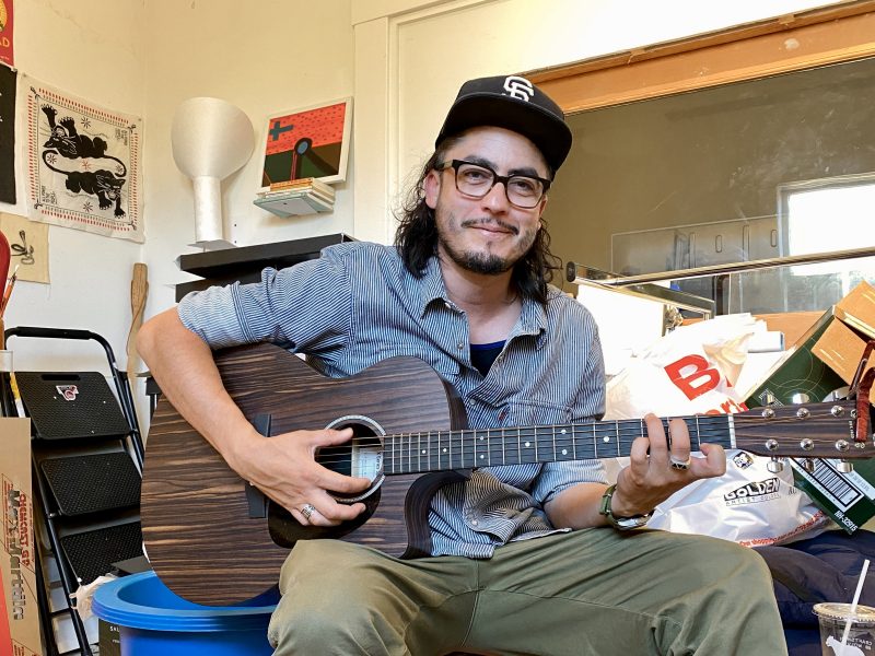 A man wearing glasses and a baseball cap sits indoors, playing an acoustic guitar. Various household items and artwork are visible in the background.