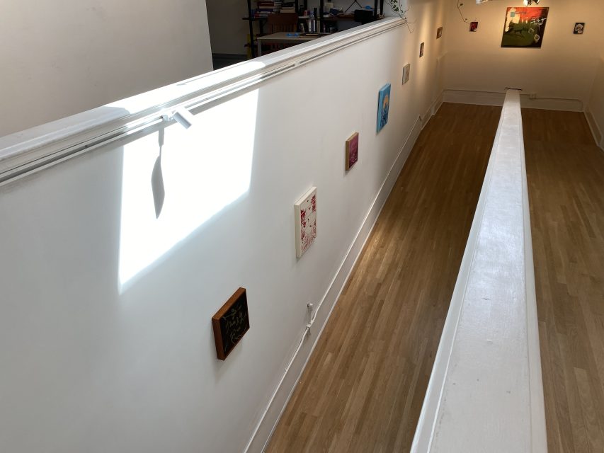A hallway with wooden flooring and white walls displays small framed artworks; sunlight shines through a window, casting a shadow on the wall.