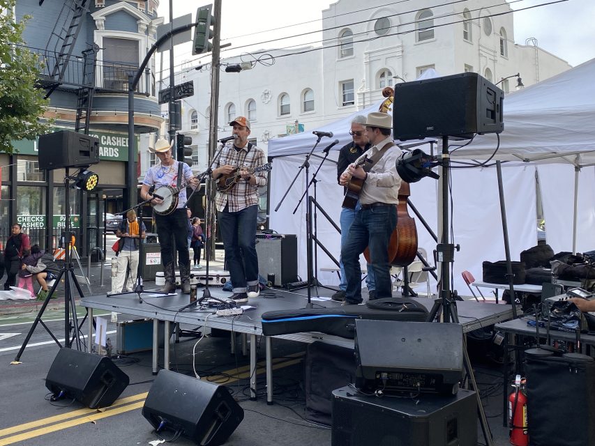 A four-member band performs on an outdoor stage with instruments including a banjo, guitar, and upright bass; buildings and street signs are visible in the background.