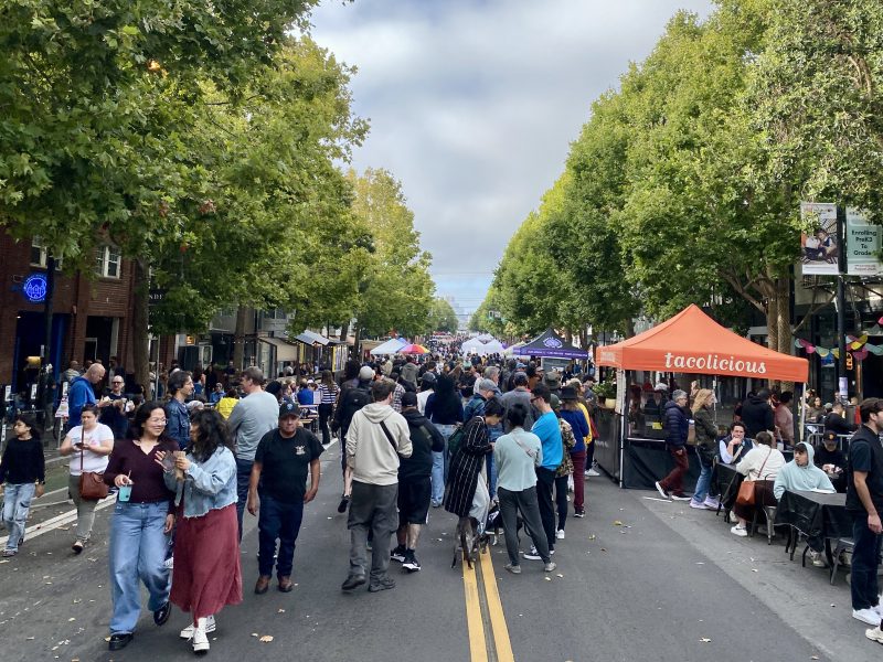 A large crowd walks down a tree-lined street at an outdoor market or festival, with vendor tents and people eating at tables on both sides.