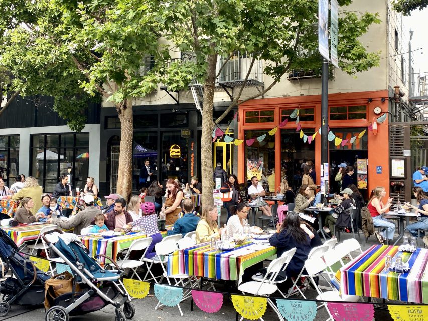 People sit at tables with colorful striped tablecloths on a city street, decorated with papel picado banners and bunting, outside shops and under trees during a community event.
