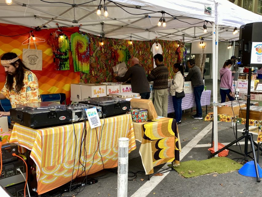 Outdoor market stall with tie-dye clothing and art, DJ equipment on table, and several people browsing under a white canopy.