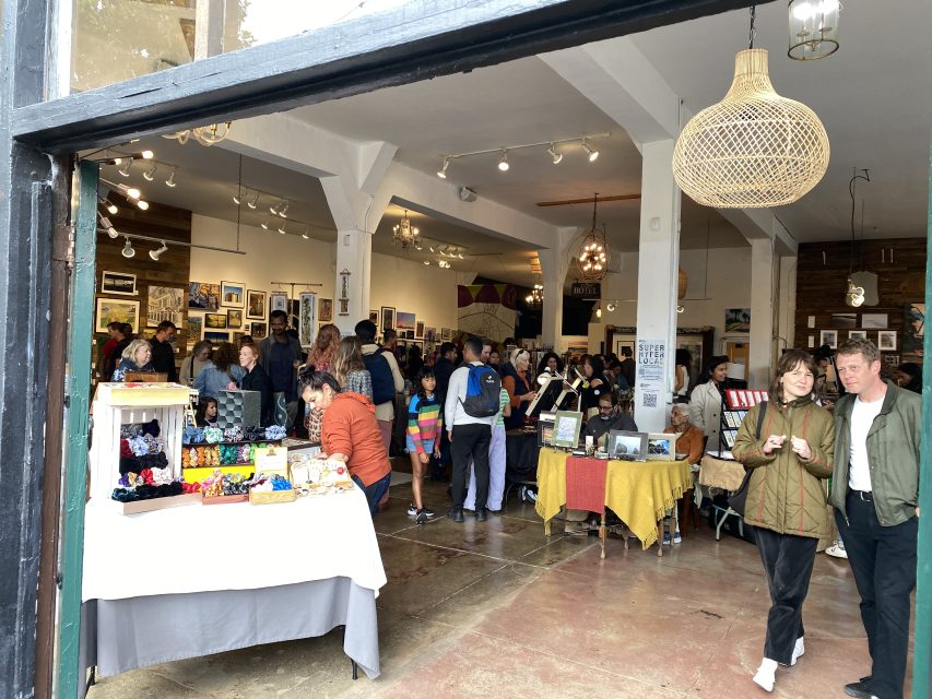 People browse art and craft booths inside a brightly lit gallery space, with various artworks displayed on the walls and tables.