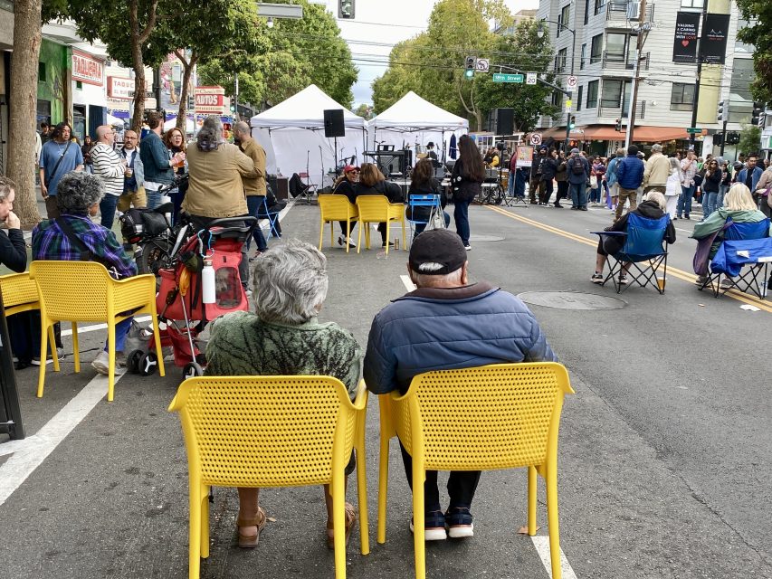 People sit on yellow chairs facing a street stage at an outdoor event; others stand or walk nearby on a closed-off city street lined with buildings and trees.