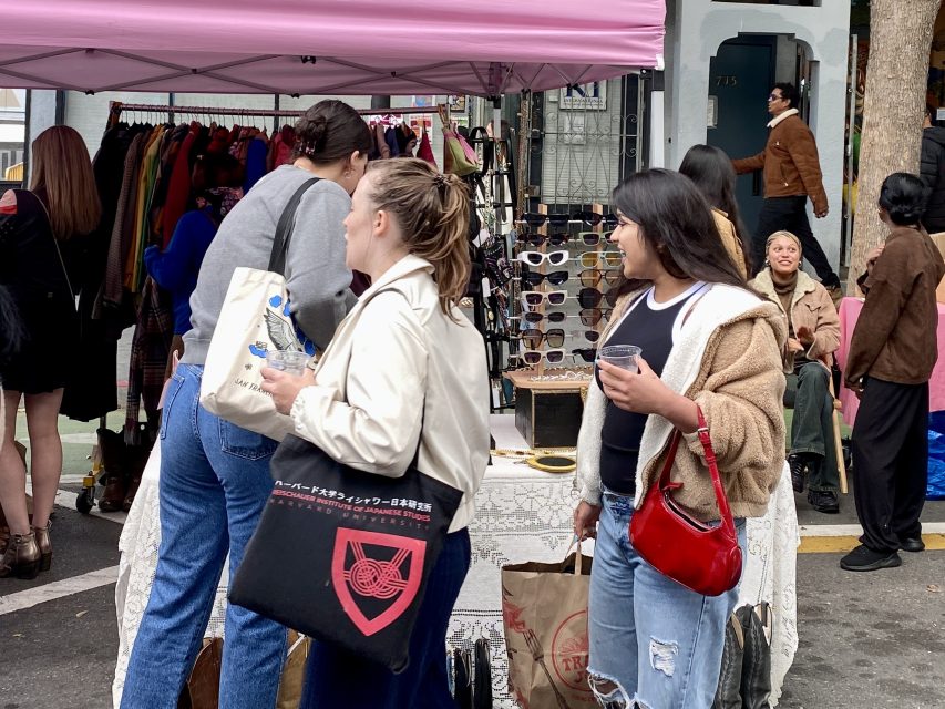 People browse clothes and sunglasses at an outdoor market stall under a pink canopy. Two women in the foreground carry tote bags and one holds a drink.