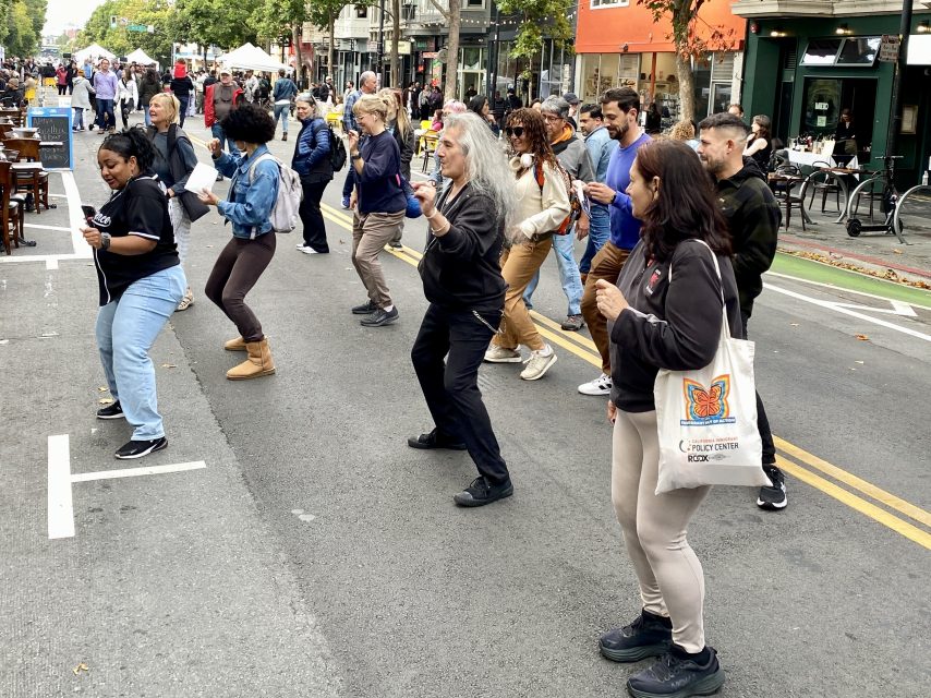 A group of people participate in a group dance on a city street during an outdoor event, surrounded by shops and trees.