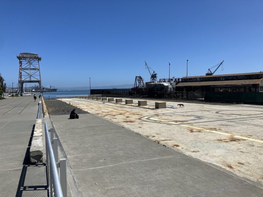 A waterfront industrial area with cranes, a dog sitting on the left walkway, and another dog walking near the center under a clear blue sky.