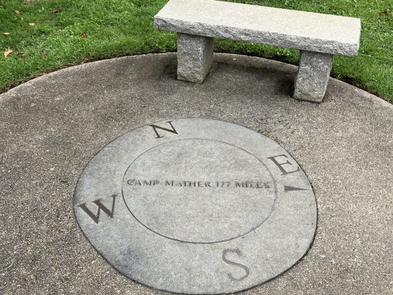 Stone bench beside a circular compass rose on the ground, marked with cardinal directions and "Camp Mather 177 Miles" engraved in the center. Grass surrounds the area.