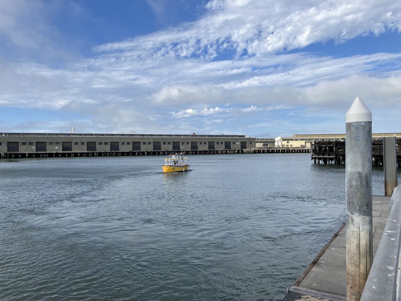 A small yellow boat travels on calm water near a dock, with a large building and partly cloudy sky in the background.
