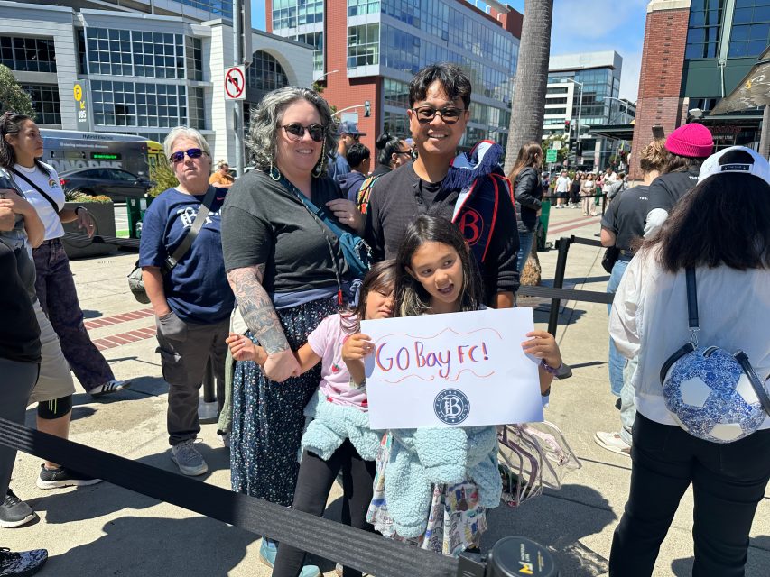 A family stands outside among a crowd. Two children hold a handmade sign that reads, “GO BAY FC!” on a sunny day.