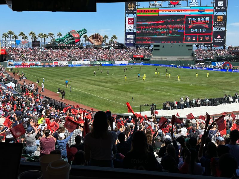 A large crowd cheers at a daytime soccer match in a stadium, with players on the field and a scoreboard displaying game details in the background.