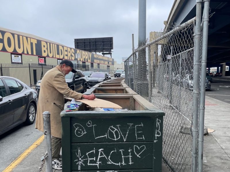 A person in a coat stands by a green dumpster with "LOVE EACH OTHER" written in chalk, next to a chain-link fence and a street under an overcast sky.