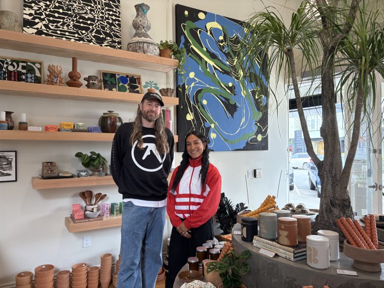 Two people stand side by side inside a shop filled with pottery, plants, and art on the walls, with shelves and a large potted tree visible.