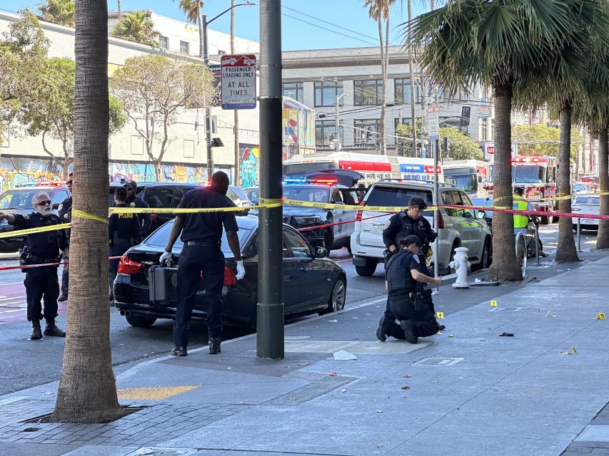 Several police officers work at a taped-off city street scene with evidence markers, police cars, and emergency vehicles visible in the background.