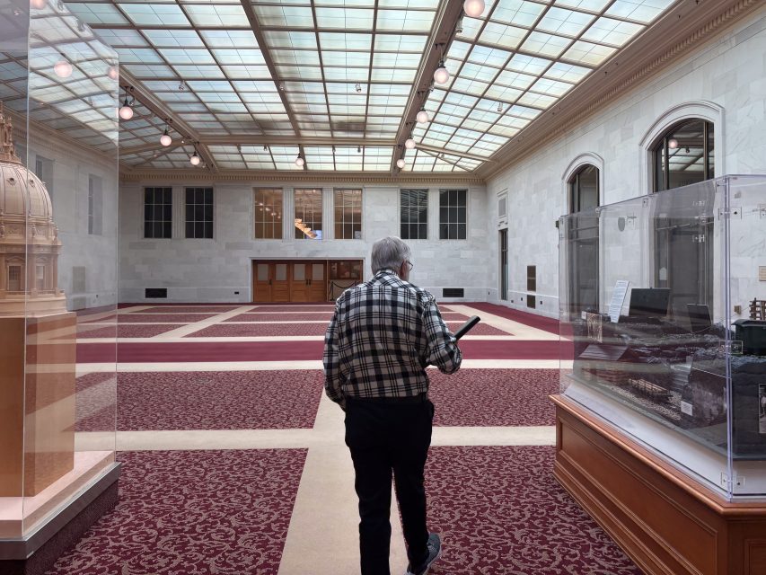 An older man in a plaid shirt walks through a spacious, carpeted hall with glass display cases and a large skylight ceiling.