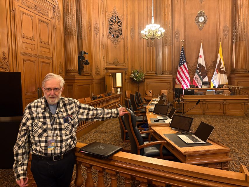 An older man in a plaid shirt stands in a wood-paneled council chamber with several empty chairs, laptops, and flags in the background.