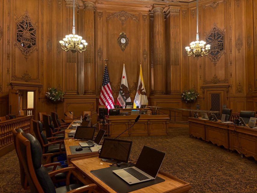 A government chamber with wood-paneled walls, several laptops on desks, flags behind the main desk, chandeliers, and ornate decor.