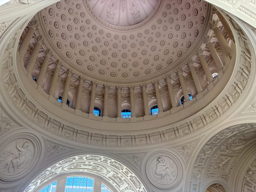Interior view of an ornate domed ceiling with detailed carvings, round windows, and classical architectural elements in a historic building.
