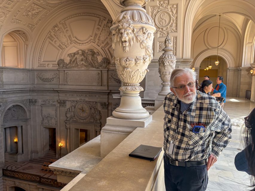 An older man in a plaid shirt stands by a stone railing in an ornate building with classical architectural details and sculptures.