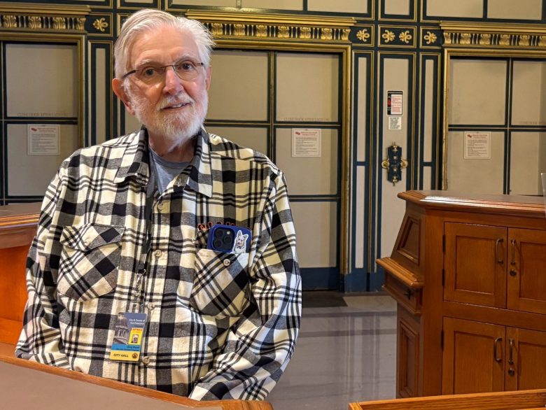 An older man in a plaid shirt sits behind a wooden desk at San Francisco City Hall; a sign advertises docent tours and provides contact information.