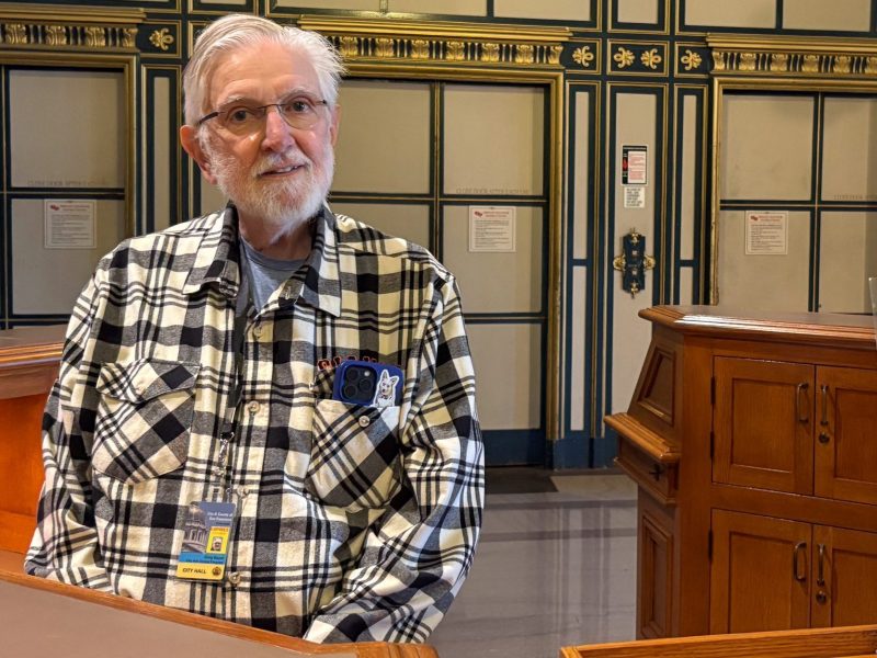 An older man in a plaid shirt sits behind a wooden desk at San Francisco City Hall; a sign advertises docent tours and provides contact information.