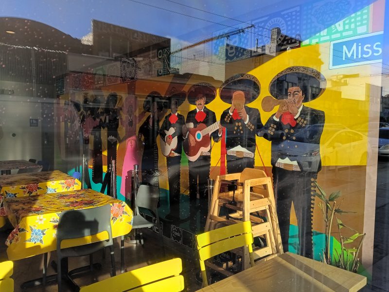 A mural of mariachi musicians decorates the interior wall of a restaurant with colorful tablecloths and empty yellow chairs, seen through a sunlit window.