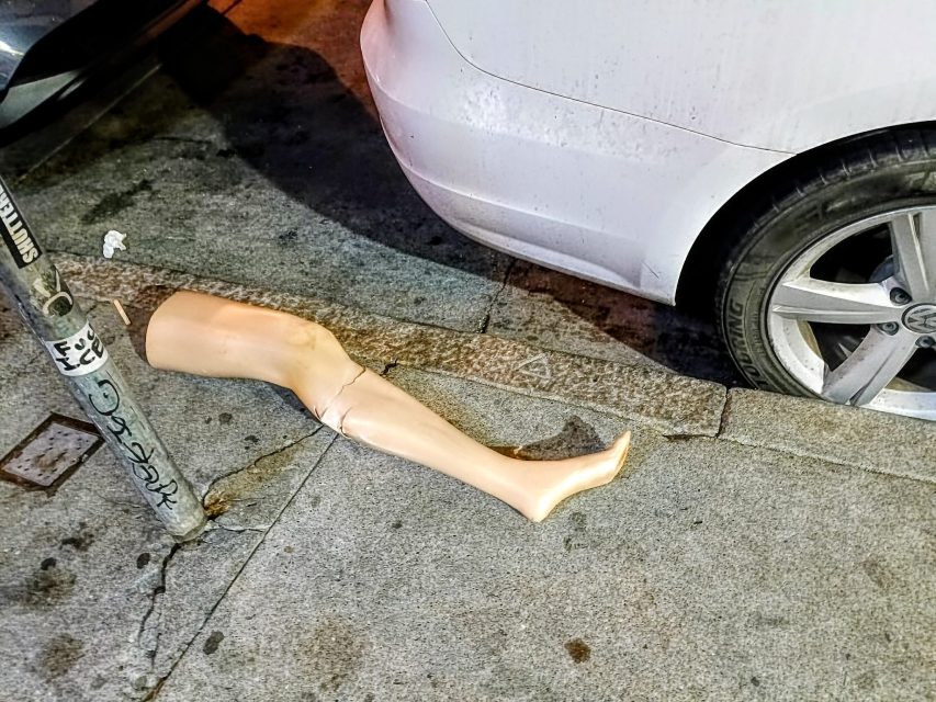 A broken mannequin leg lies on a city sidewalk next to a parked white car and a metal pole.