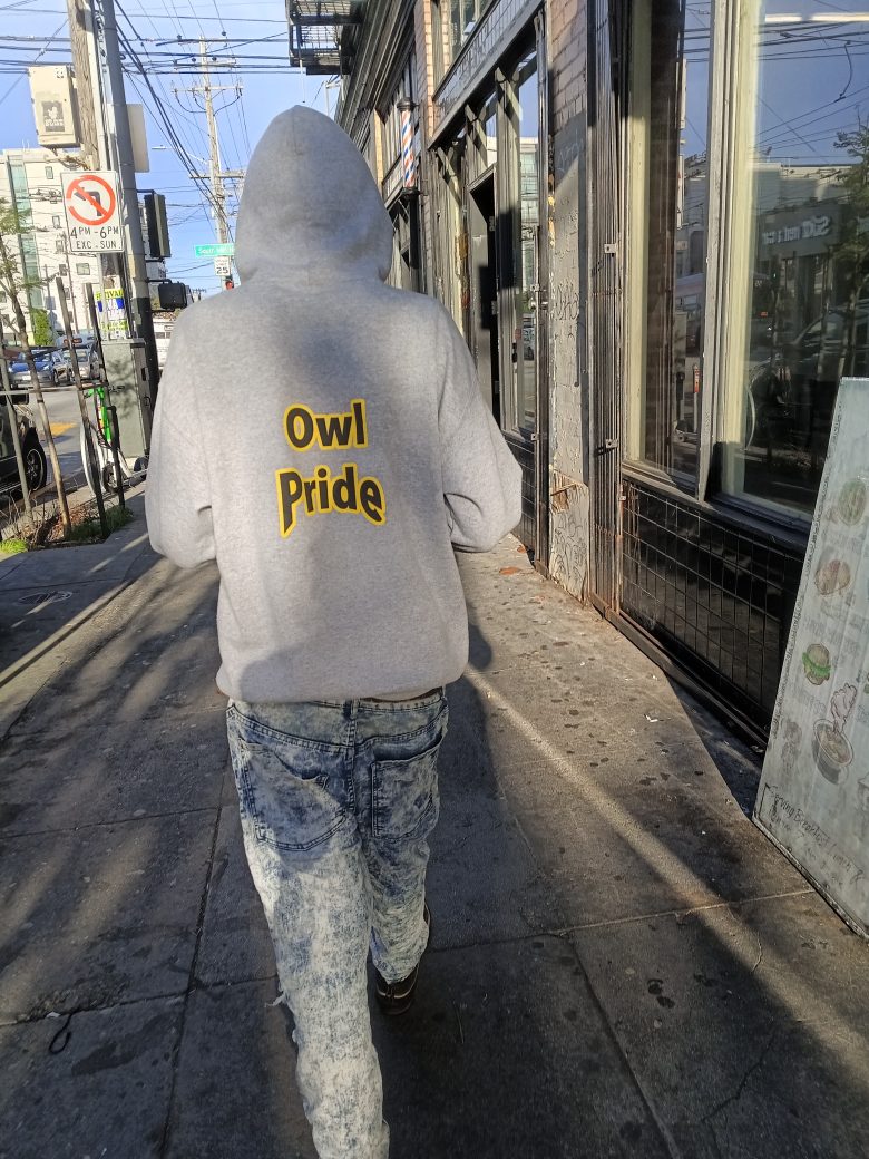 Person wearing a gray "Owl Pride" hoodie and jeans walks on a sunlit city sidewalk near storefronts and street signs.