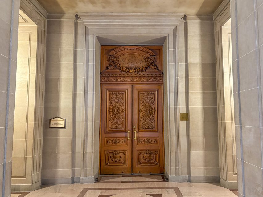 Ornate double wooden doors with intricate carvings, set in a stone wall. Two plaques are mounted on either side of the doorway in a formal, well-lit hall.