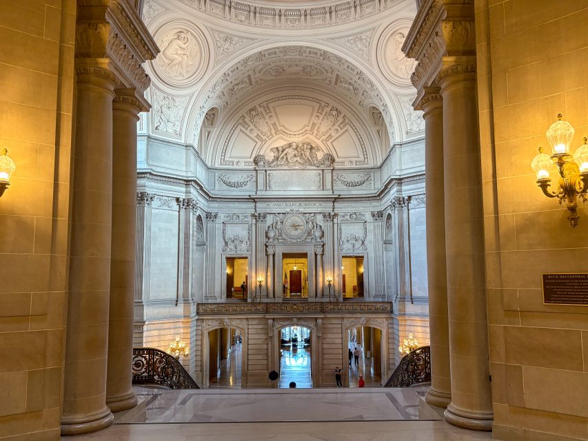 Grand interior hall with ornate columns, detailed carvings on the arched ceiling, large windows, and people walking on marble floors below.
