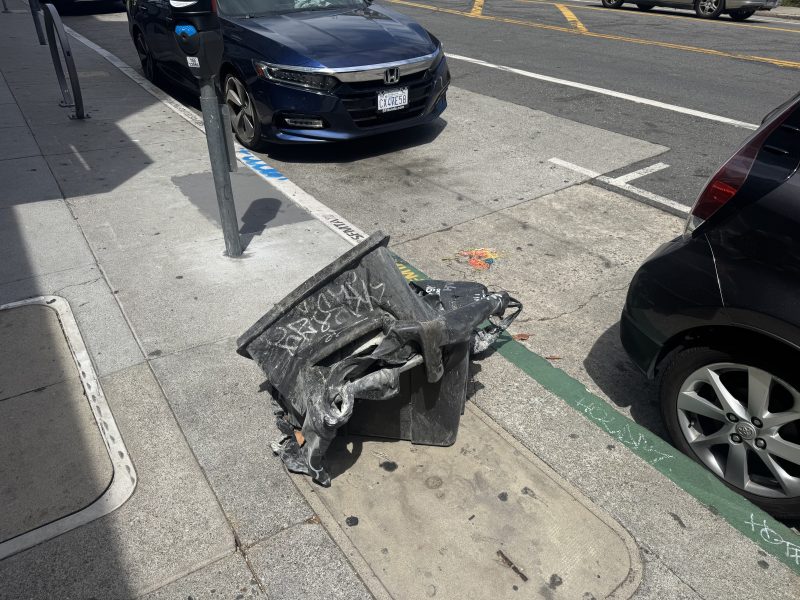 A damaged and overturned trash bin with graffiti is lying on a city sidewalk next to parked cars.
