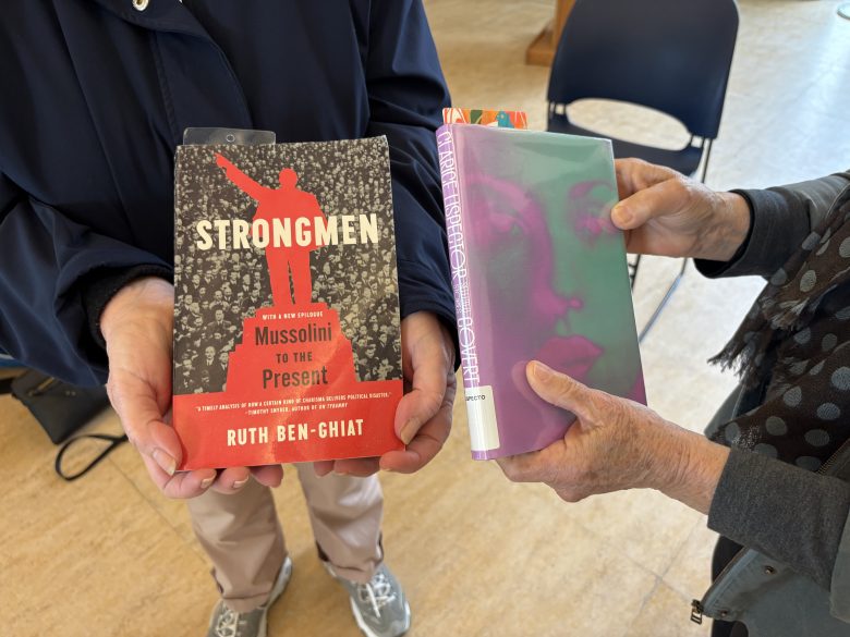 Two people each hold a book: one holds "Strongmen: Mussolini to the Present" by Ruth Ben-Ghiat, the other holds a book with a green-tinted cover featuring a woman's face.