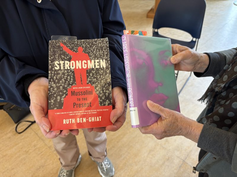 Two people each hold a book: one holds "Strongmen: Mussolini to the Present" by Ruth Ben-Ghiat, the other holds a book with a green-tinted cover featuring a woman's face.