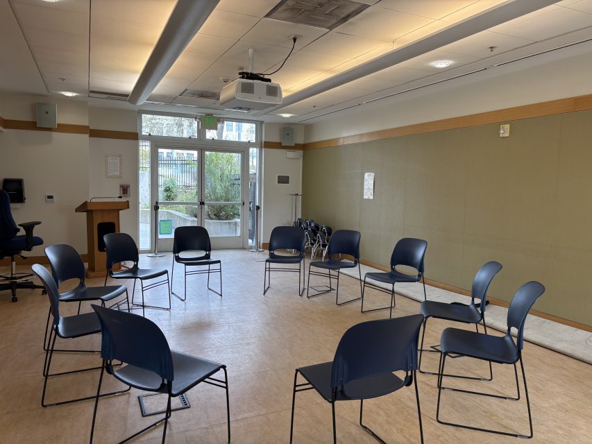 A circle of empty chairs is arranged in a well-lit meeting room with a podium, projector, and large windows facing outside.