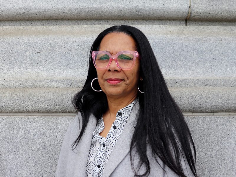 A woman with long black hair, wearing pink glasses, hoop earrings, a patterned shirt, and a light coat stands in front of a stone wall, her poised expression hinting at a quiet strength amid whispers of scandal.