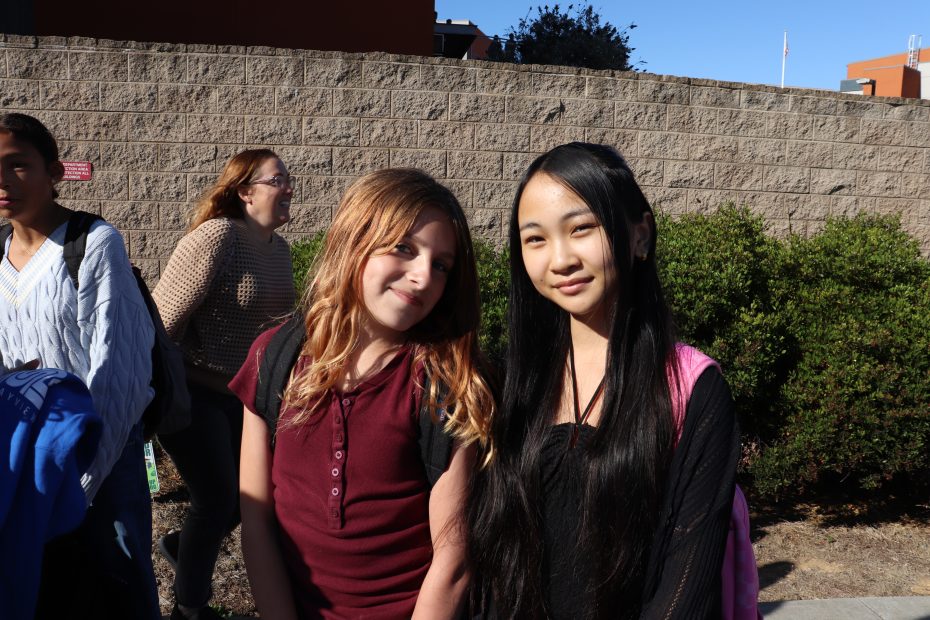 Two girls with backpacks stand outside in sunlight, smiling at the camera, with two other people and a brick wall in the background.